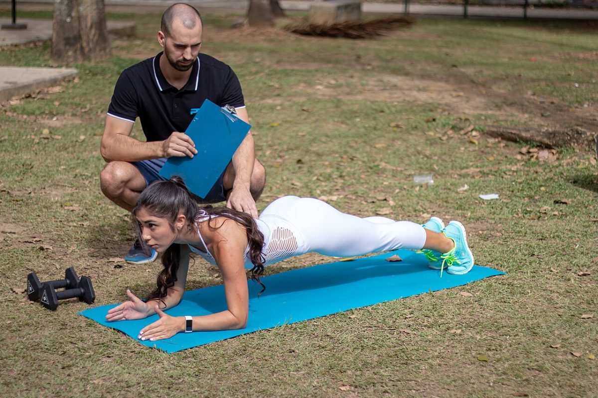 Mujer haciendo una plancha sobre los antebrazos en una esterilla, con un entrenador al lado.