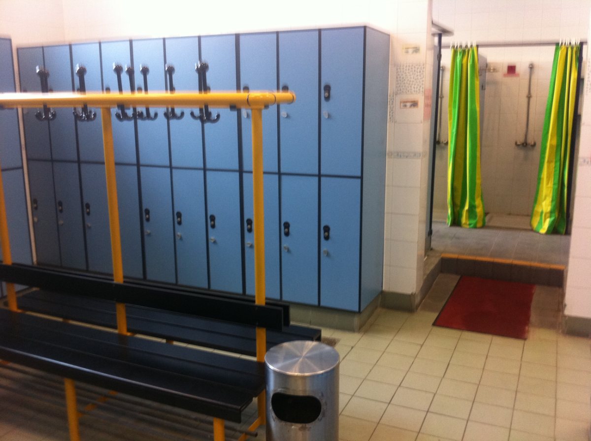 Vestuario de un polideportivo con taquillas, banco y duchas al fondo.