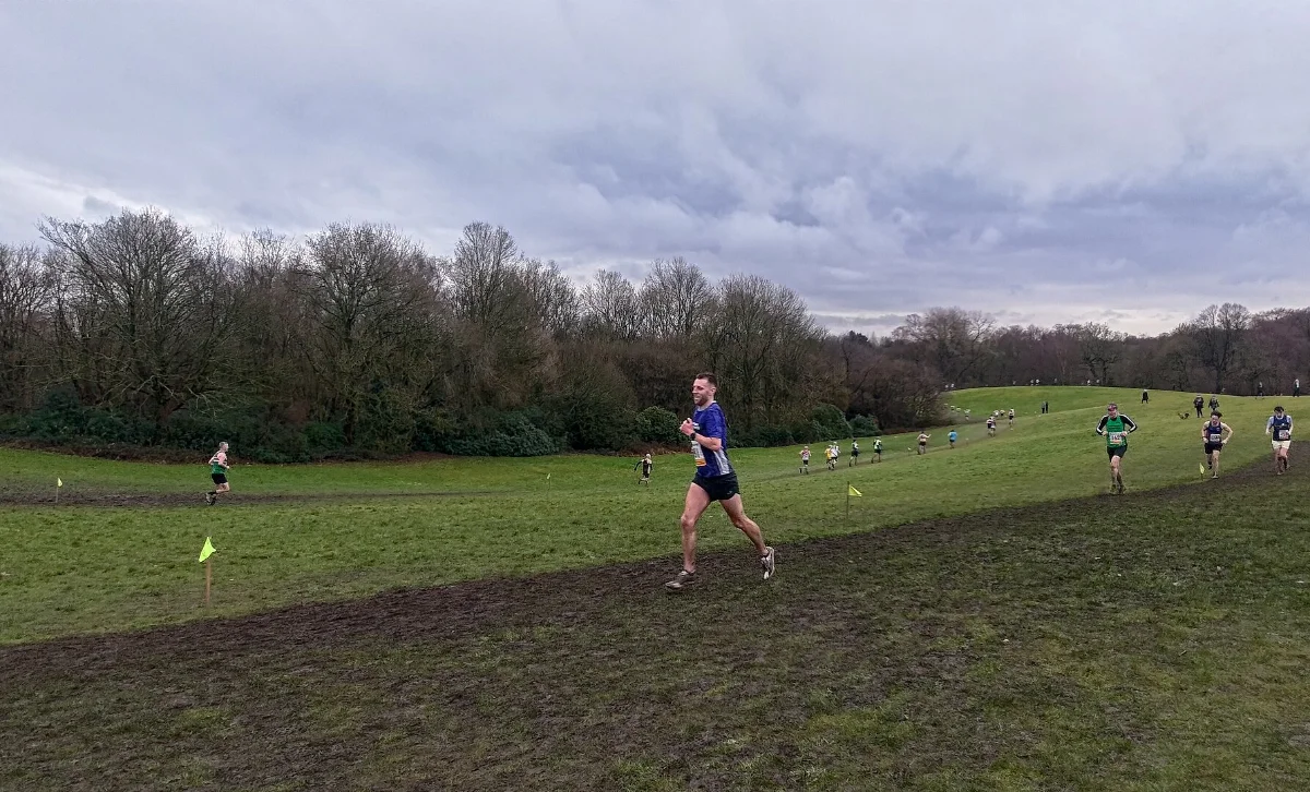 Corredores en una carrera de cross por una pradera con barro, con árboles al fondo.