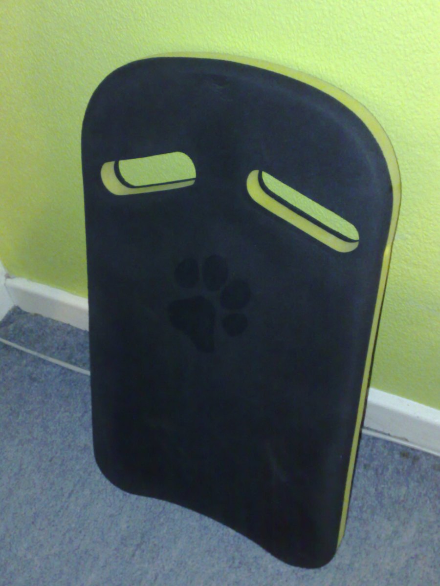 Tabla de natación de espuma negra con dos huecos para las manos, apoyada en una pared.