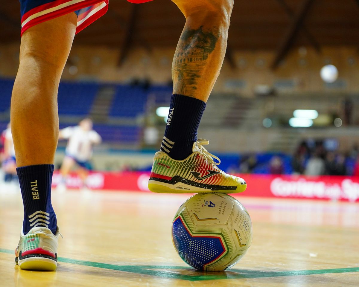 Jugador de fútbol sala con el pie sobre el balón en una pista de parquet, con zapatillas de suela lisa y calcetines con agarre